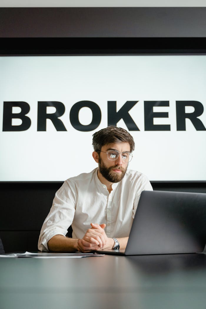 Young businessman working on a laptop in a modern office with a 'BROKER' sign.